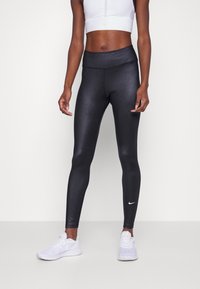 Černé legíny s vysokým pasem, hladkým a lesklým povrchem a malým bílým logem Nike blízko kotníku, nošené s bílými sportovními botami.
