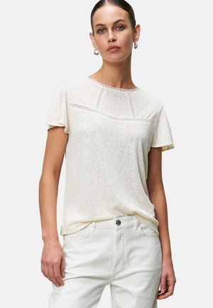 Cream short-sleeve top with a round neckline, featuring decorative stitching across the chest. Paired with light-colored, fitted pants.