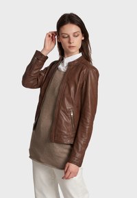 Brown leather jacket with a fitted design, standing collar, and zippered pockets. Worn over a beige sweater and white pants.