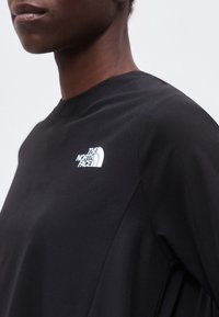 Άτομο με μαύρο μακρυμάνικο μπλουζάκι με το λογότυπο The North Face στο στήθος, εμφανίζεται από τον ώμο έως το πηγούνι μπροστά σε μια απλή φόντο.