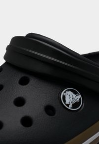 Εγγύς λήψη μαύρου περφορέ κλογκ με ρυθμιζόμενο λουράκι στη φτέρνα και κυκλικό λογότυπο με καρικατούρα κροκοδείλου.