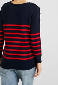 Marineblauer Strickpullover mit roten horizontalen Streifen, Rundhalsausschnitt und Knopfdetails an der Schulter. Gerippte Bündchen und Saum. Lässige Passform.