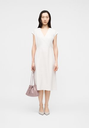 Femme aux longs cheveux foncés portant une robe midi blanche sans manches et des talons pointus de couleur claire, tenant un petit sac à cordon mauve.