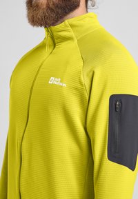 Giacca gialla brillante con un motivo a texture e righe e una tasca con zip nera sul braccio sinistro. Presenta un colletto alto e una zip parziale.