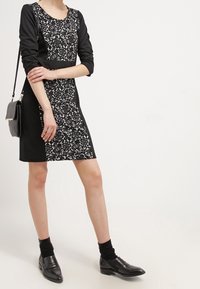 Svart och vit klänning med en spetsmönstrad överdel och enfärgade svarta ärmar. Har en rak silhuett och knälång klippning. Bärs med svarta skor.