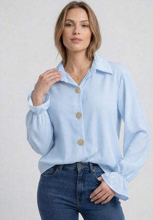 Kobieta w jasnoniebieskiej bluzce z dużymi ozdobnymi guzikami i bufkami, w połączeniu z ciemnoniebieskimi dżinsami, na jednolitym tle.