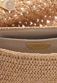 Gewebte Strohtasche in hellem Beige mit einer strukturierten Oberfläche, ausgestattet mit einem Beigen Innenfutter aus Stoff und einem goldfarbenen Markenschild im Inneren.