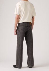Homme portant un jean en denim gris foncé à coupe ample et un t-shirt blanc à manches courtes, vu de dos sur un fond uni.