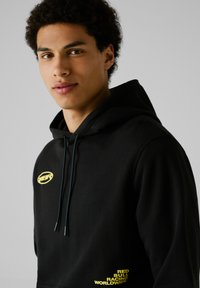 Sudadera con capucha negra y un bolsillo tipo canguro en el frente. Presenta un logo amarillo en el pecho y el texto "RED BULL RACING WORLDWIDE" en el lateral.