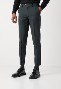 Op maat gemaakte donkergroene broeken van textiel met een slanke pasvorm, zijzakken en een enkele knoopsluiting. Gecombineerd met zwarte schoenen.