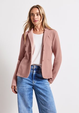 Blazer en tissu texturé rose doux avec des revers, des boutons sur le devant et deux poches latérales, associé à un T-shirt blanc et un jean bleu clair.