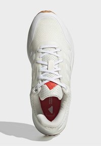 Chaussure de sport blanche avec une tige en mesh texturé, un col matelassé, un accent rouge sur la semelle intérieure et des lacets blancs sur une coque d'orteil renforcée.