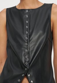 Haut sans manches noir en similicuir avec une rangée centrale de boutons-pression métalliques argentés, partiellement défait en bas.