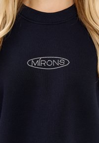 Granatowy sweatshirt z prążkowanym dekoltem, z okrągłym logo "MRONS" ozdobionym kryształkami na piersi. Gładka tekstura, luźny krój.