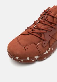 Sneaker in suede marrone con superficie testurizzata, chiusura con lacci e suola in gomma caratterizzata da dettagli di spruzzi bianchi. Punta rotonda e elementi di design sobri.