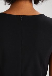 Černé šaty s hladkým, matným povrchem mají kulatý výstřih a skrytý zip na zádech, což vytváří bezproblémový vzhled.