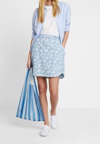 Žena v modro-páskované sukni, bílé teniskách, bílém tričku a světlemodré košili, držící velkou modro-bílou pruhovanou tašku.