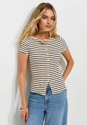 Donna con capelli biondi lunghi indossa una camicia beige a righe nere a maniche corte con bottoni e jeans blu, in piedi contro un muro bianco.