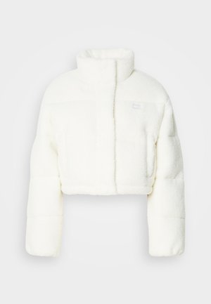 Apgriezts pūkains jaka baltā mākslīgā kažokā, ar augstu apkakli, polsterētu dizainu un mazu logotipu uz krūšu daļas. Mīksta tekstūra un strukturēta forma.