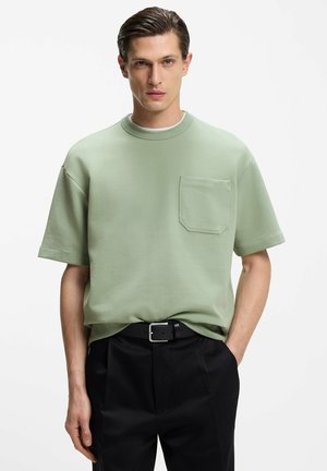 Mies, joka pukeutunut vaaleanvihreään lyhythihaiseen paitaan, jossa on rintatasku, ja mustiin housuihin, seisoo toisessa kädessään taskussa yksinkertaisen taustan edessä.