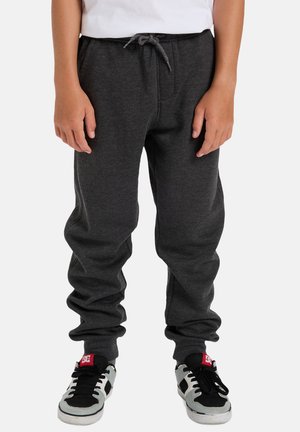 Grijze katoenen trainingbroek met een elastische tailleband en trekkoord, voorzien van zijzakken en een taps toelopend ontwerp. Gepaard met zwarte en grijze sneakers.