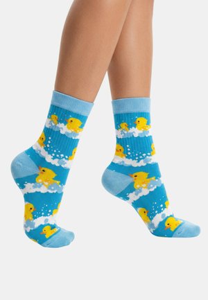 Siniset sukat, joissa keltaisia kumipullloja, jalassa valkoista taustaa vasten.