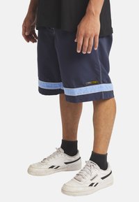 Granatowe szorty do pływania z jasnoniebieskimi pasami, naszywką z logo i luźnym krojem, zestawione z białymi i czarnymi sneakersami oraz czarnymi skarpetkami.