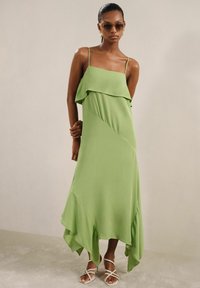 Rochie maxi verde fluidă, cu volane suprapuse la bust, hem asimetric și bretele subțiri. Pur și simplu combinată cu sandale albe cu curea.
