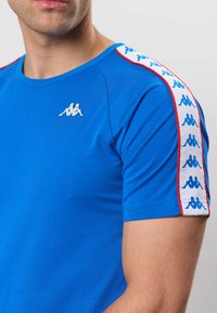 Mees, kes kannab kuninglikus sinises T-särgis, millel on õlal valge ja sinine logoriba ning rinnal väike valge logo, lihtsa tausta ees.