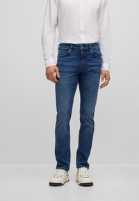 Blå denimjeans med slim passform, fem fickor och midjehöjd. Bärs med en vit skjorta med knappar och crèmefärgade sneakers.