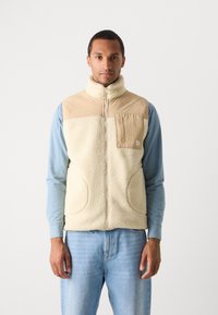 Fleece vest in cream with a tan nylon upper section and pocket, featuring a high collar and front zip, paired with a light blue shirt.