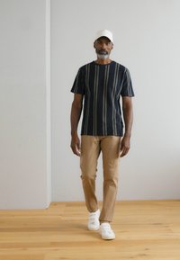 Vīriešu svītrainā krekls tumši zils ar bēšām vertikālām līnijām, apvienots ar brūniem biksēm un baltām kedām. Modelis valkā baltu cepuri. Gluda tekstūra.