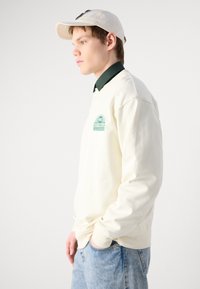 Felpa color crema con un logo verde sul petto, indossata sopra una camicia colletto verde scuro, abbinata a jeans blu chiaro.