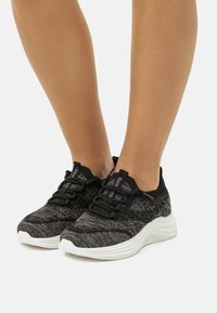 Zapatillas de punto negras y grises con un acabado texturizado, que cuentan con una suela blanca acolchada, cordones negros y acentos morados en la lengüeta.