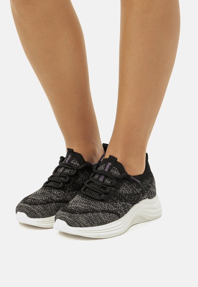Zapatillas de punto negras y grises con un acabado texturizado, que cuentan con una suela blanca acolchada, cordones negros y acentos morados en la lengüeta.