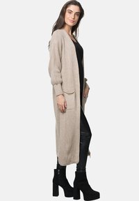Beige stickad cardigan med långa ärmar, öppen front och sidofickor. Bärs över en svart topp och byxor i konstläder, ihop med svarta klackboots.