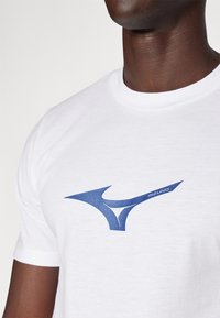 Balta kokvilnas T-kreklā ar zilu Mizuno logo. Kreklam ir apaļa kakla izgriezums un īsas piedurknes, kā arī gluda tekstūra.