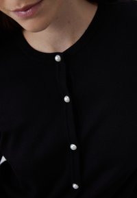 Cardigan nero con colletto rotondo e cinque bottoni di perla lungo il davanti, realizzato in tessuto a maglia liscia. Texture chiara e dettagli dei bottoni visibili.