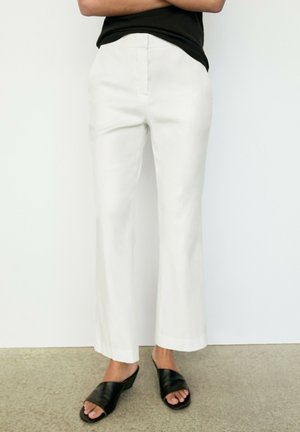 Person trägt weiße gerade geschnittene Hose und schwarze offene Slip-On-Schuhe und steht auf einem hellen Boden vor einer weißen Wand.