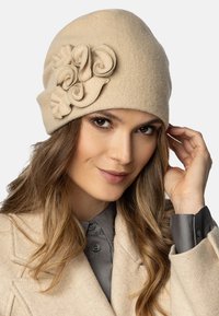 Chapeau en laine beige avec un accent floral, présentant des détails en tissu roulé. Texture douce et forme arrondie ajustée conçue pour la chaleur.