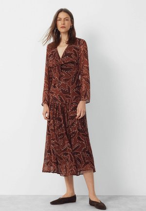 Kobieta ubrana w brązową sukienkę midi z długim rękawem, ozdobioną abstrakcyjnymi wzorami liści, oraz płaskie ciemnobrązowe buty, stojąca na białym tle.