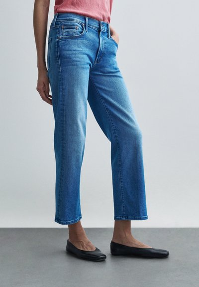 Lyseblå denimjeans med lige pasform, fem lommer og diskrete whiskering-effekter, kombineret med flade sorte sko.