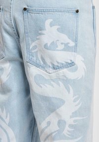Jeans de mezclilla azul claro con un gran diseño de dragón blanco impreso alrededor del bolsillo trasero y la zona de la pierna.