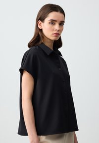 Blusa nera a maniche corte con colletto, realizzata in tessuto morbido. Presenta una chiusura frontale con bottoni e un design dal taglio rilassato.