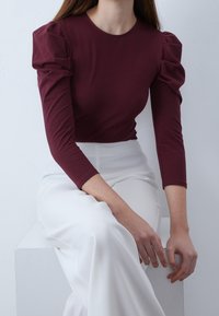 Burgundy åtsittande topp med puffärmar, i kombination med vita, flödande byxor. Slät tyg med subtil textur, placerad mot en ljus bakgrund.