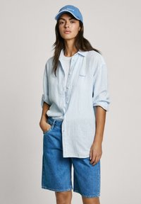 Ein hellblau gestreiftes Hemd über einem weißen T-Shirt, kombiniert mit blauen Jeans-Bermudashorts. Eine blaue Mütze rundet den lässigen Look ab.