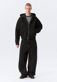 Jeune homme portant un sweat à capuche noir zippé, une chemise rayée, un pantalon large noir et des chaussures noires, debout avec une main dans la poche.