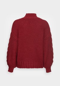 Maglione rosso lavorato a maglia con colletto alto, motivo testurizzato, maniche ampie e orlo a costine, che mette in mostra una costruzione in filato spesso.