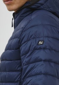 Námornícka modrá prešívaná bunda s kociek, ktorá má čiernu nášivku s logom na rukáve a vysoký golier pre väčšie teplo.