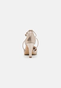 Chaussure à talon aigu dorée en métal avec un bout pointu, une bride de cheville et un talon stiletto élancé. Texture lisse avec une finition brillante.